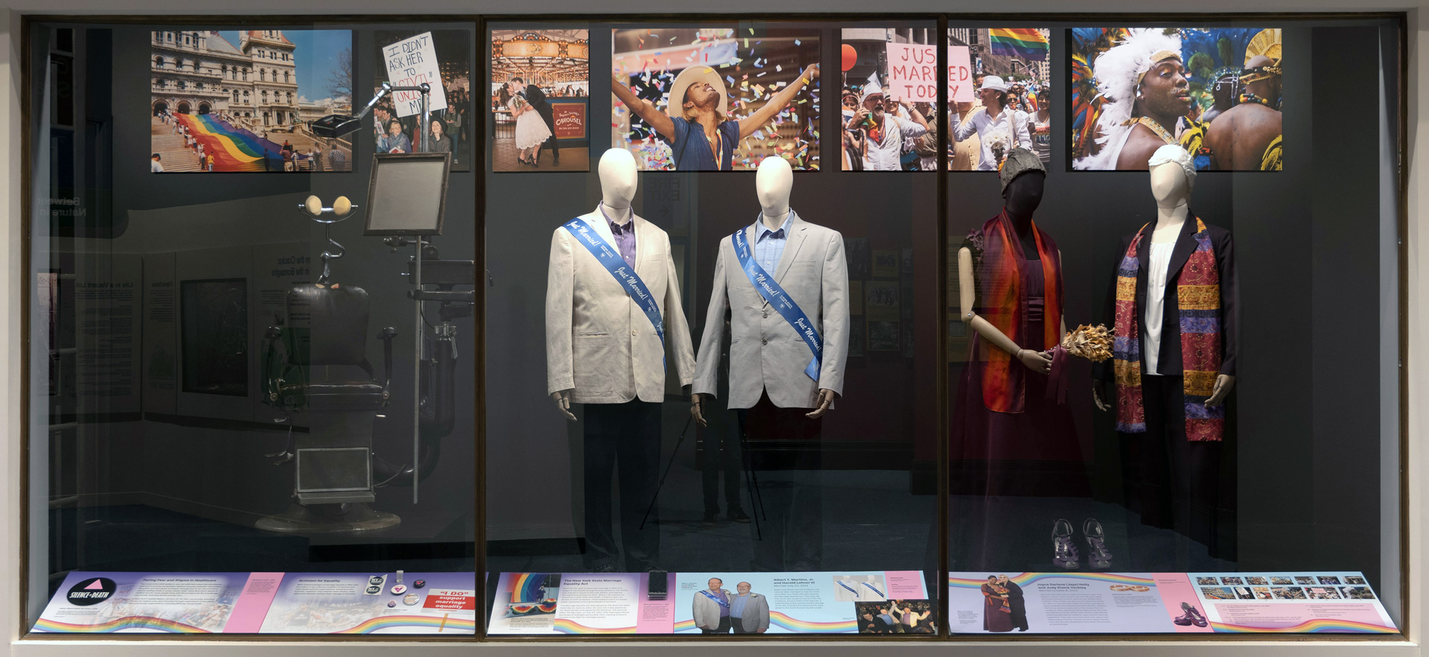 view of exhibit case displaying a vintage opthalmalogy chair, two men's suits with Just Married sashes, and two women's outfits