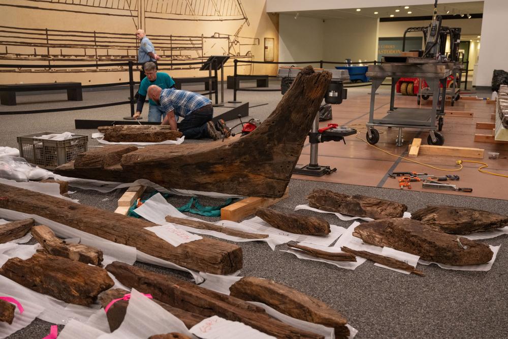 Two staff members oversee a room full of large wooden pieces from a disassembled boat