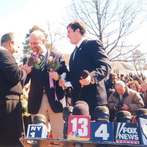 Two men exchange vows while a third man stands by with a microphone infront of a large crowd. In the foreground are microphones from different news agencies.