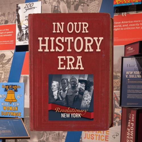 In Our History Era title text on a red book cover over the main graphics for the exhibition Revolutionary New York and a background photograph from the gallery space