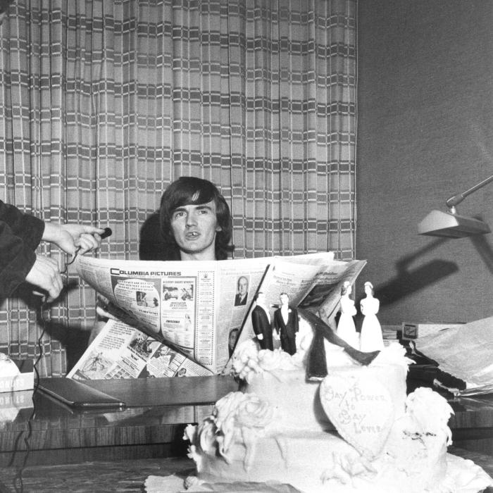 Black/white photograph of a man seated at a desk, holding an open newspaper. A figure at the far left holds a small microphone towards the seated man. In the foreground on the desk is a wedding cake with toppers featuring same sex couples.