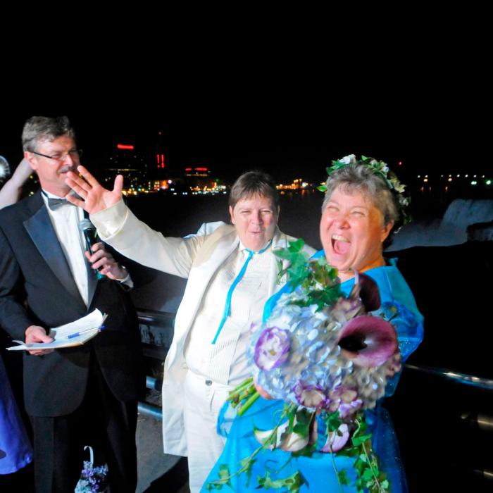 Two women cheer and smile after exchanging vows in front of Niagara Falls. At left stand a wedding officiant and wedding party members.