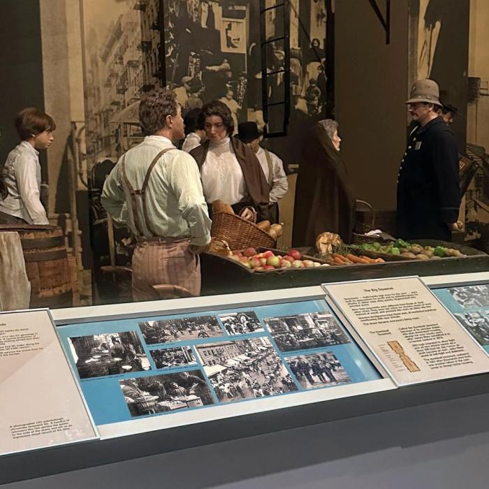 Gallery of 19th-century mannequins at a market behind a text/image panel